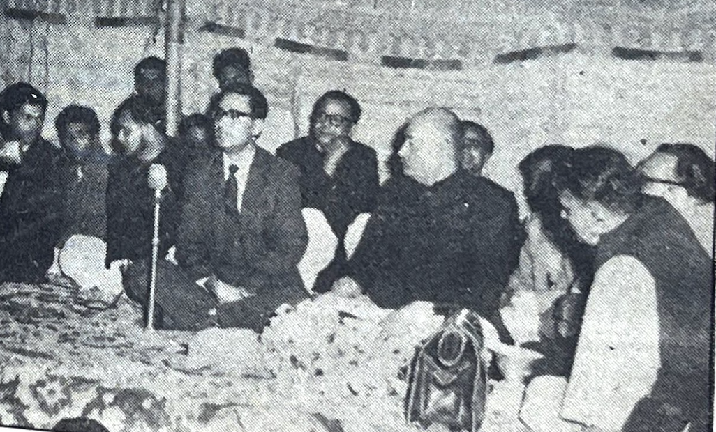 Black and white image showing a group of men sitting on a stage and a man in a suit in front of a microphone